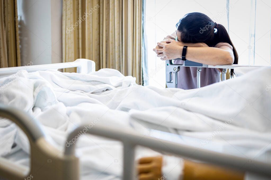 Tired asian woman fell asleep at the beside a patient in a hospital bed,physical fatigue caused by overwork or stress of caring for the sick senior,caregiver taking a nap with head down on crossed arm