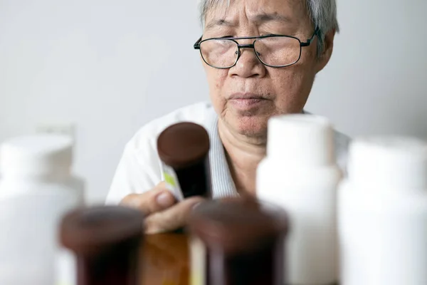 Asian senior woman holding drug bottle in front of medicine cabinet in her home,reading the label on a bottle for medical treatment,old elderly checking expiration date or deterioration of medication