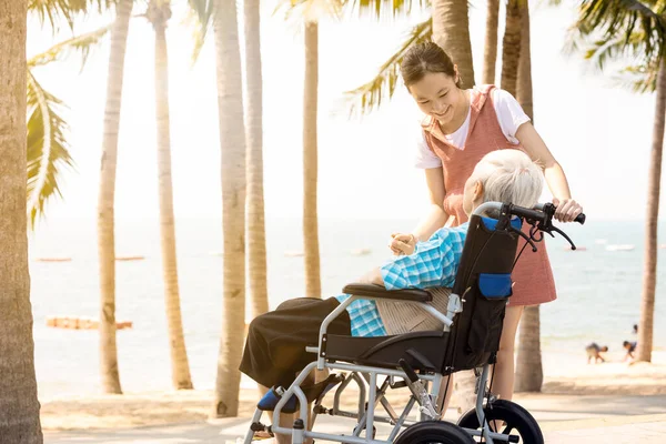 Happy asian old elderly in a wheelchair granddaughter relaxing enjoying on the sea beach together,teenager girl show love,take care support her senior grandmother while traveling in summer vacation