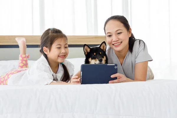 Happy Asian mother and daughter lying on bed and using digital tablet. Black Shiba Inu on bed between its owner. Concept family with dog at home.