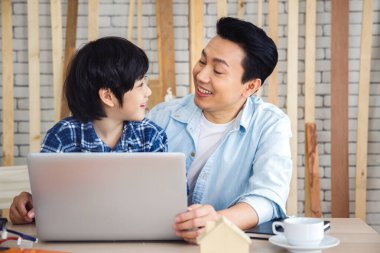 Mutlu Asyalı baba ve oğul dizüstü bilgisayar kullanıyor ve marangozluk atölyesinde birbirlerini arıyorlar. Aile kavramı evde birlikte hobi yapıyor..