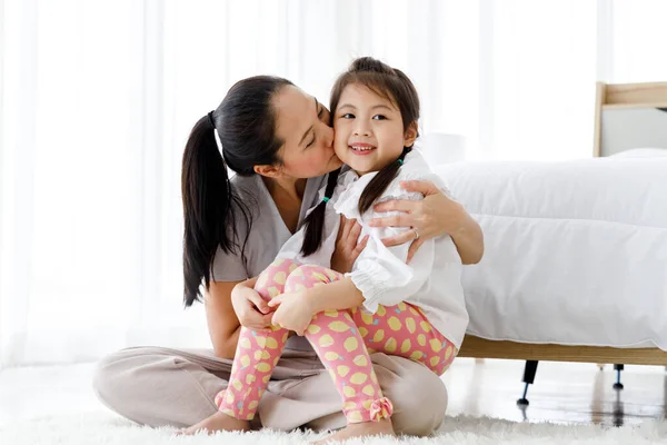 Asian mother kissing her daughter in bright bedroom at home.
