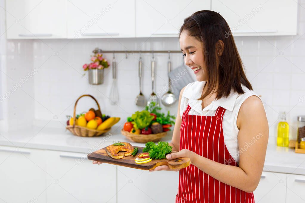 Asian woman chef in red apron holding salmon steak, serving on wooden board. Concept woman preparing meals at home.