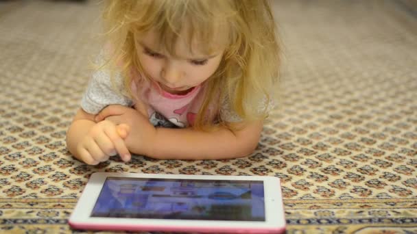 Linda niña trabaja en la tableta de ordenador acostado en la alfombra — Vídeo de stock