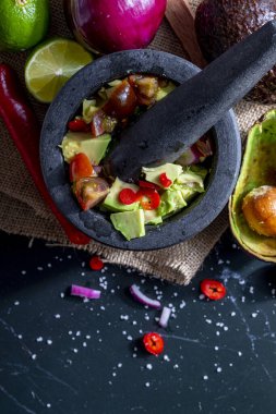 Geleneksel bir taş havanda avokado, limon, soğan, domates ve kırmızı biber gibi tüm malzemeleriyle guacamole hazırlamak. İştah açıcı ve sağlıklı geleneksel guacamole. Ev yapımı görünüş. Üst Manzara. Kopyalama alanı ile.