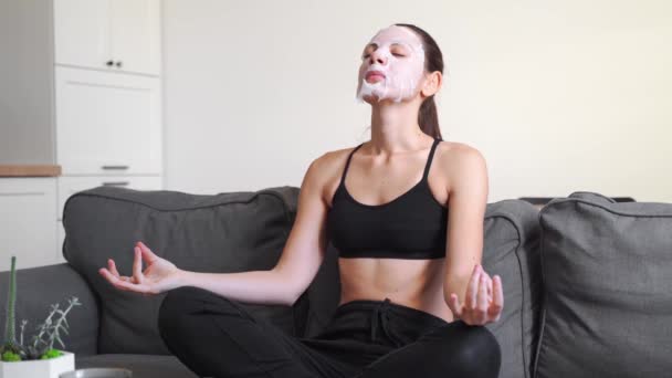 Jovem mulher milenar em casa aplicando máscara de pano no rosto e meditando. Bem-estar tempo relaxante — Vídeo de Stock