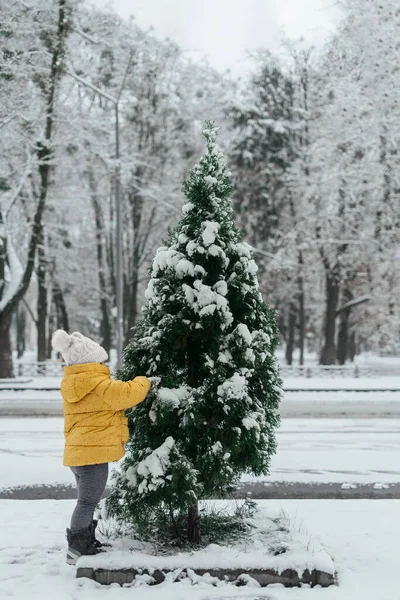 Niño pequeño en traje amarillo en el día nevado afuera con pino cubierto de nieve — Foto de Stock