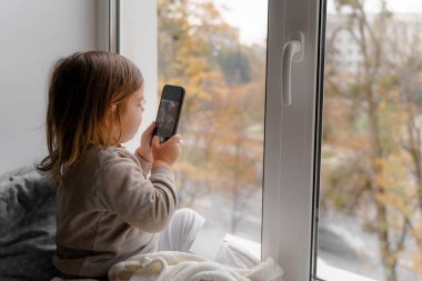 Küçük çocuk evde telefon kullanıyor, fotoğraf çekiyor ve oyun oynuyor.