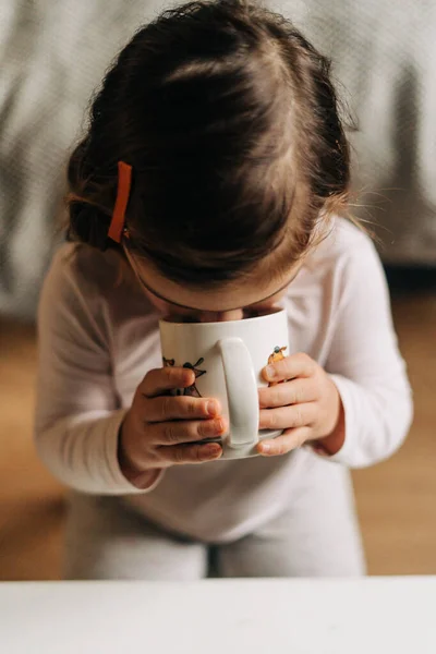 Белая малышка пьет какао из кружки дома. Рождественские огни — стоковое фото