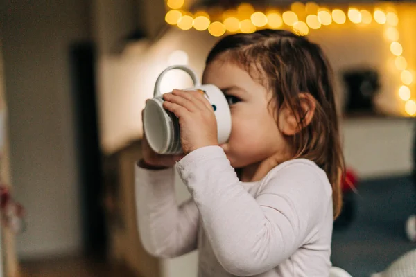 Menina branca bebê bebendo cacau de caneca em casa. Natal bokeh luzes — Fotografia de Stock