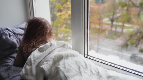 Barnet ligger på fönstret i vardagsrummet och tittar på gatan. mysigt hem på helger, helgdagar och inlåsning — Stockvideo