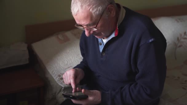 Hombre mayor sentado en casa y usando un teléfono inteligente. Entrega en línea. Medicina virtual. Sitio web de noticias. Conexión con la familia — Vídeo de stock