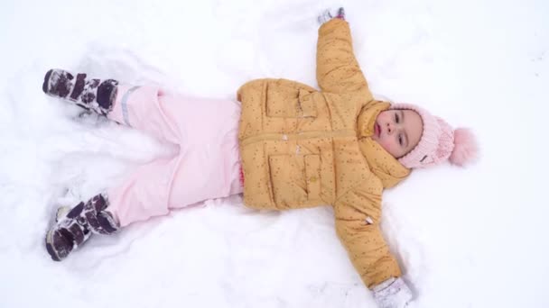 Niña en ropa de invierno haciendo ángel de nieve en un día de invierno. Gran actividad al aire libre con los niños. Una vida franca. Feliz infancia. — Vídeo de stock