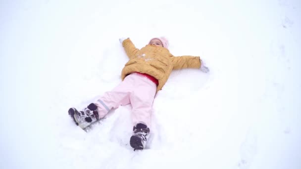 Peutermeisje in winterkleding die sneeuwengel maakt op een winterdag. Buitenactiviteiten met kinderen. Echt eerlijk leven. Gelukkige jeugd — Stockvideo