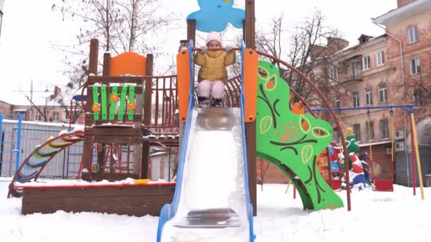 冬の遊び場で遊んでいる幼児の女の子。スイングで雪の日。雪と子供の冬の活動。子供のための寒い気候の雪のスーツ. — ストック動画