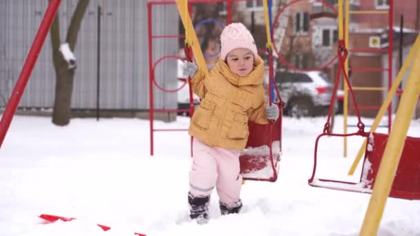 Pikkulapsi tyttö leikkii leikkikentällä talvella. Luminen päivä vauhdissa. Lapset talvella toimintaa lunta. Kylmä sää lumipuku lapselle. — kuvapankkivideo