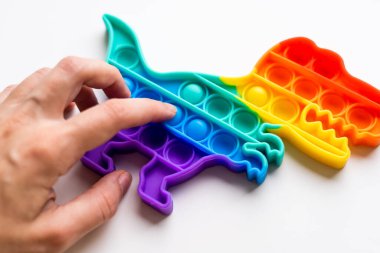 Gökkuşağı renginde ve dino şeklinde Popit gökkuşağı oyuncağı. Moda baloncuklu pop müziği. Stres önleyici tuhaf bir eğilim. Beyaz arkaplan ve parmak kabarcıkları patlatıyor.