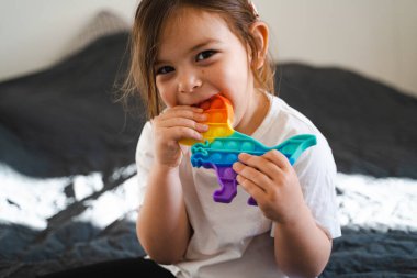 Gökkuşağı renginde popit oyuncağı olan küçük bir kız. Stres atmak için dino şeklinde silikon oyuncak. Kabarcıklar duyusal modayı takip ediyor. Çocuk evde samimi bir şekilde oynuyor