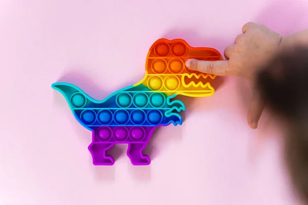 Popit juguete arco iris en colores arcoíris y forma de dino. moda burbuja pop fidget. Divertida tendencia sensorial anti estrés. fondo rosa y el dedo haciendo estallar las burbujas. — Foto de Stock