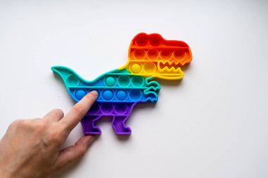 Gökkuşağı renginde ve dino şeklinde Popit gökkuşağı oyuncağı. Moda baloncuklu pop müziği. Stres önleyici tuhaf bir eğilim. Beyaz arkaplan ve parmak kabarcıkları patlatıyor.