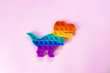 Gökkuşağı renginde ve dino şeklinde Popit gökkuşağı oyuncağı. Moda baloncuklu pop müziği. Stres önleyici tuhaf bir eğilim. pembe arkaplan ve parmak kabarcıkları patlatıyor.