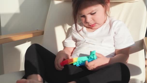 Дівчина Тоддлер з поп-іграшкою в веселкових кольорах. Силіконова іграшка для зняття стресу у формі динозавра. Бульбашки чутливі модні стрімкі. Дитина грає вдома у відвертому стилі — стокове відео