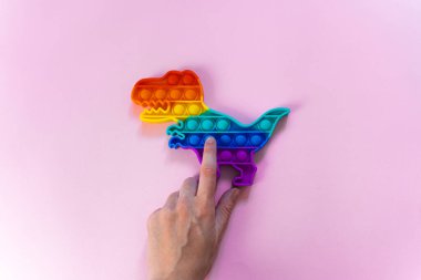 Gökkuşağı renginde ve dino şeklinde Popit gökkuşağı oyuncağı. Moda baloncuklu pop müziği. Stres önleyici tuhaf bir eğilim. pembe arkaplan ve parmak kabarcıkları patlatıyor.