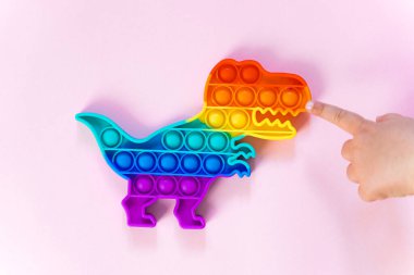 Gökkuşağı renginde ve dino şeklinde Popit gökkuşağı oyuncağı. Moda baloncuklu pop müziği. Stres önleyici tuhaf bir eğilim. pembe arkaplan ve parmak kabarcıkları patlatıyor.