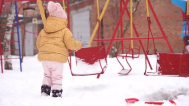Küçük kız kışın çocuk parkında oynuyor. Salıncakta karlı bir gün. Çocuklar kar ile kış aktivitesi. Çocuk için soğuk hava elbisesi.