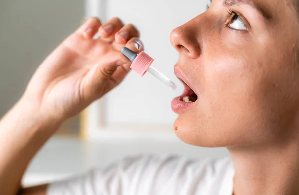 Mujer sosteniendo un aceite de cannabis sublingual pipeta gotero y dejándolo caer en su boca. Mujer joven enferma o enferma que usa una gota para medicarse — Foto de Stock