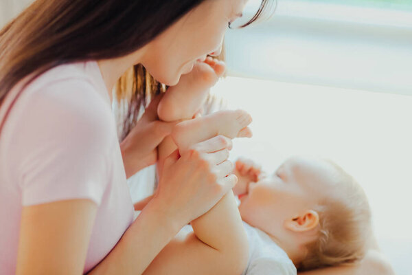 Молодая счастливая мать целует ноги своего малыша. Уход за ребенком и материнство