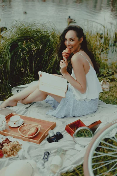 Beautiful playful woman is reading book and eating croissant at picnic. Lifestyle, outdoor recreation on weekends