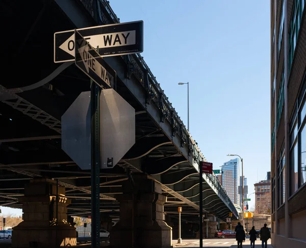 Two \'One Way\' street signs intersecting under a bridge in dramatic light with silhouettes of pedestrians in the background in New York City