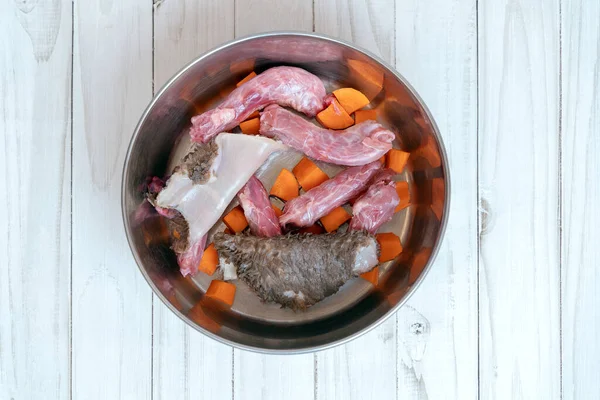 Top view on metal pet bowl with chicken necks, beef paunch pieces and cut carrot. The part of natural organic daily meal ration preparation for dogs concept. Copy space, white wooden table background.