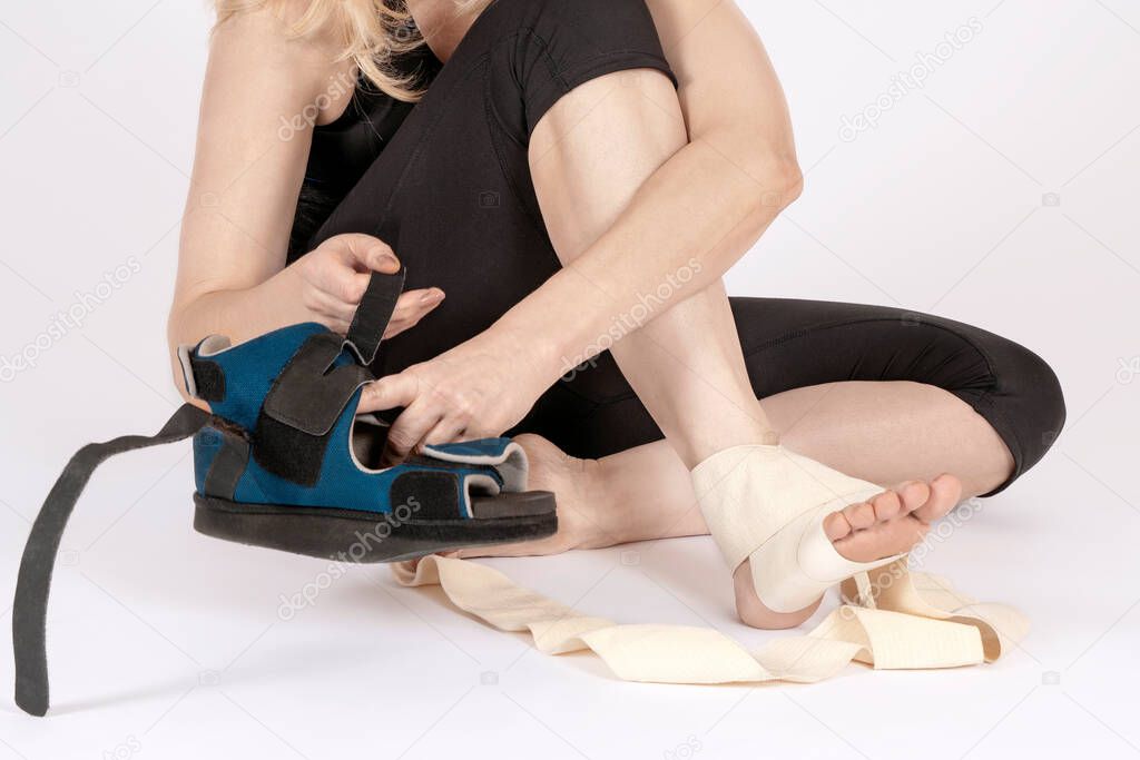 Female wearing sport clothes, sitting on the floor, bandages her leg and takes up the orthopedic boot. Recovery after trauma or surgery.  White background, copy space.