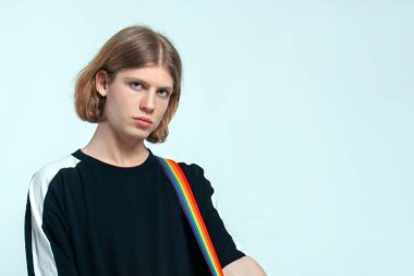 Sarışın, yeşil gözlü, omzunda gökkuşağı şeridi olan yakışıklı genç adam kameraya bakıyor. Sıradan siyah. Hafif arkaplan, yumuşak seçici odak, kopyalama alanı.