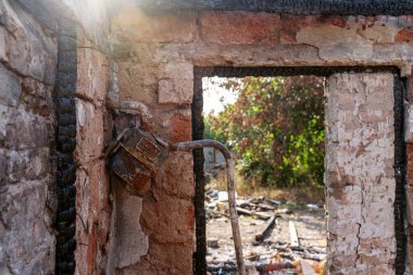 Ev yangınla yok oldu. Kayıp pencere, hasarlı gaz sayacı, yanmış ahşap çerçeve, çatlak duvarlar, harabeler. Güneş çatıdaki delikte parlıyor. 
