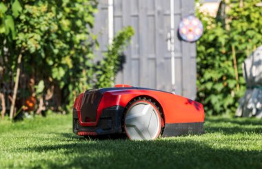 Otonom kırmızı robotik çim biçme makinesi barakanın yanındaki güneşli bahçede çim biçiyor. Çim bakımı için modern akıllı bahçe malzemeleri. Arkaplan, metin için yer, kopyalama alanı.