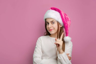 Pembe Noel Baba şapkalı ve pembe arka planda beyaz gömlekli gülümseyen kızın portresi. Noel kutlaması ve kış tatili konsepti.