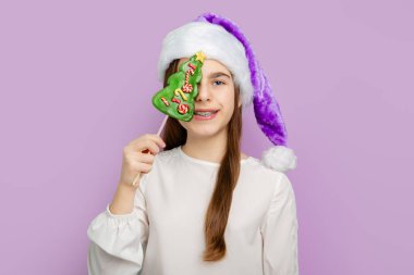 Noel Şapkalı Kız Ağaç Şekeri Tutuyor. Mor Noel Baba şapkalı neşeli kız pastel arka planda Noel ağacı şeklinde şekerler tutuyor..