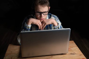 Dizüstü bilgisayarda gözlüklü ve tişörtlü çocuk öğreniyor ve düşünüyor.