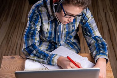 Tişörtlü ve gözlüklü çocuk dolmakalemle dizüstü bilgisayara not alıyor.