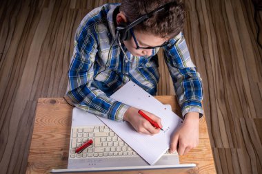 Tişörtlü ve gözlüklü çocuk dolmakalemle dizüstü bilgisayara not alıyor.