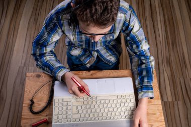 Tişörtlü ve gözlüklü çocuk dolmakalemle dizüstü bilgisayara not alıyor.