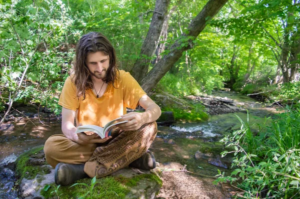Νεαρός διαβάζει ένα βιβλίο στην όχθη ενός ποταμού. — Φωτογραφία Αρχείου