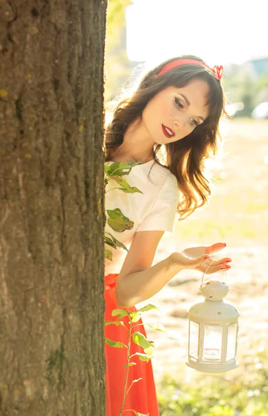 Beautiful girl looks out from behind a tree and holding a lanter — Stock Photo, Image