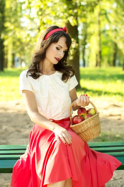 Beautiful girl sitting on a bench and holding a basket with appl — Stock Photo, Image