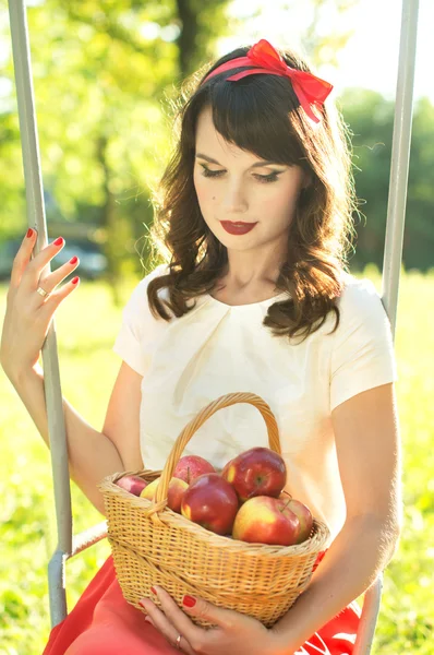 Happy girl sitting on the swing and holding a basket with apples — Stock Photo, Image