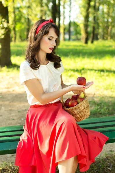 Chica feliz sentada en un banco y sosteniendo una canasta con manzanas — Foto de Stock