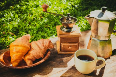 Kahve fincanı ve ahşap zeminde kruvasan ekmeği. Bahçede arka planda yeşil bir çimen var..